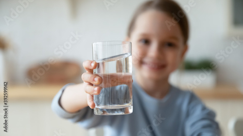 Close up smiling little girl holding glass of pure mineral water, offering to camera, cute pretty child kid recommending healthy lifestyle habit, drinking clean aqua for refreshment