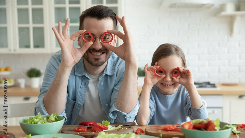 Head shot portrait smiling young father and little daughter holding red peppers circles as glasses, looking at camera, sitting at table in kitchen, posing for funny family photo, cooking together Head shot portrait smiling young father and little daughter holding red peppers circles as glasses, looking at camera, sitting at table in kitchen, posing for funny family photo, cooking together