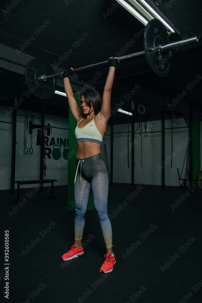 Fit young girl with light brown hair wearing black leggins, pink top and sneakers and doing squats with barbell, dark gym at background, cross training workout, portrait. Fit young girl with light brown hair wearing black leggins, pink top and sneakers and doing squats with barbell, dark gym at background, cross training workout, portrait.