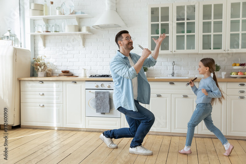 Happy young father with little daughter having fun with kitchenware, standing in modern kitchen at home, playing funny game, smiling dad holding whisk as microphone, singing, family enjoying weekend