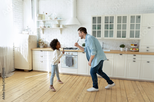Happy young father and little African American girl playing funny game, standing in kitchen, dad and adorable daughter holding kitchenware as microphones, singing, multiracial family having fun © fizkes