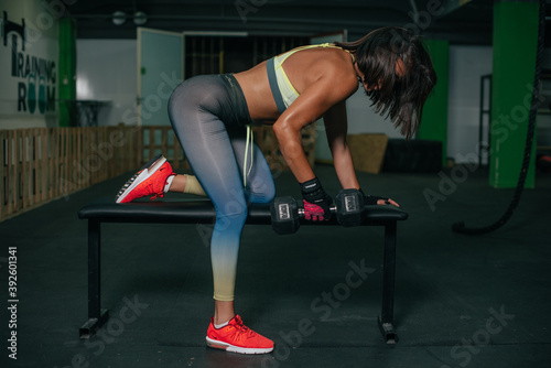 Fit young girl with light brown hair wearing black leggins, pink top and sneakers and doing squats with barbell, dark gym at background, cross training workout, portrait.