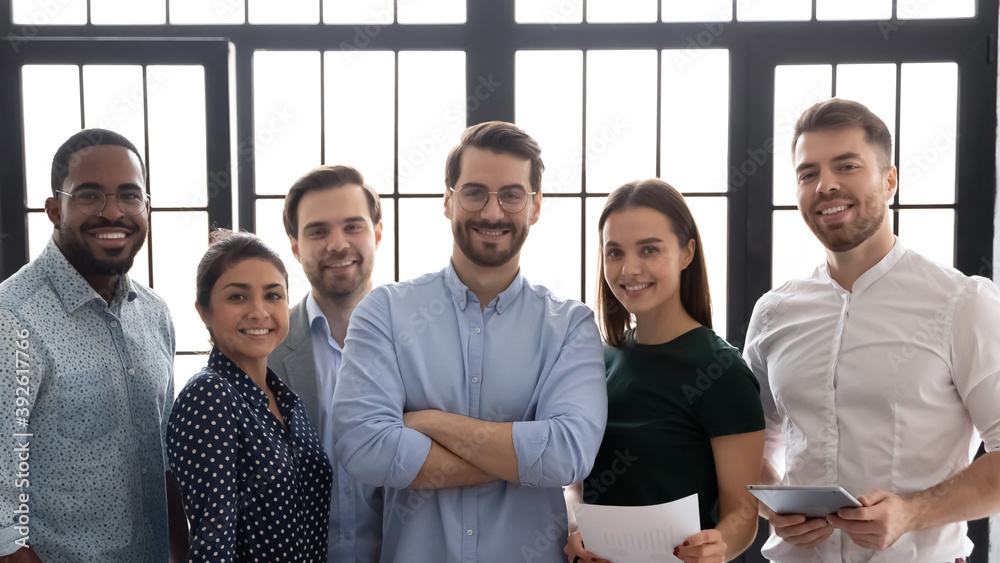 Friendly team. Group portrait of successful multiethnic business team in office, confident charismatic man leader posing indoors surrounded by happy diverse staff members smiling and looking at camera