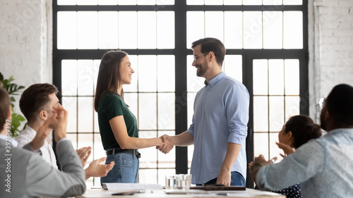 Glad millennial male leader handshaking congratulating happy female employee with professional achievement expressing gratitude for contribution in common project while diverse team staff applaud