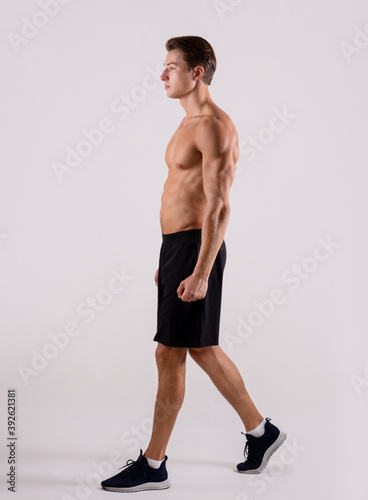 Young sporty guy with sexy bare torso walking on light studio background, side view