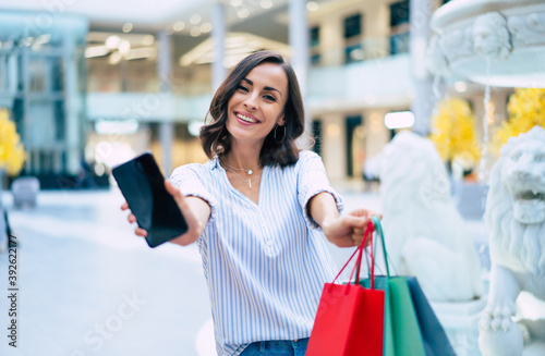 Happy beautiful young stylish woman with shopping bags is using smart phone while walking in the mall on black friday