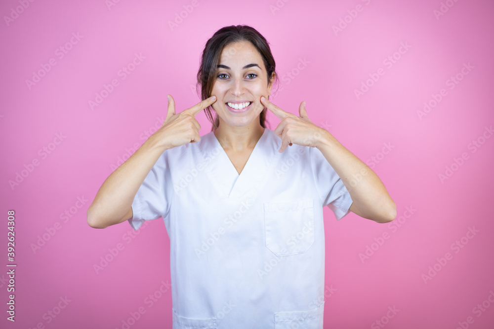 Young brunette doctor girl wearing nurse or surgeon uniform over isolated pink background smiling confident showing and pointing with fingers teeth and mouth Young brunette doctor girl wearing nurse or surgeon uniform over isolated pink background smiling confident showing and pointing with fingers teeth and mouth