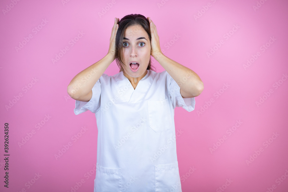 Young brunette doctor girl wearing nurse or surgeon uniform over isolated pink background crazy and scared with hands on head, afraid and surprised of shock Young brunette doctor girl wearing nurse or surgeon uniform over isolated pink background crazy and scared with hands on head, afraid and surprised of shock