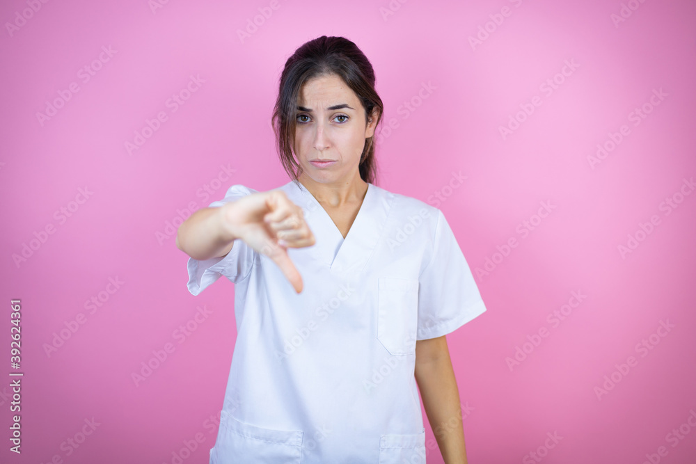 Young brunette doctor girl wearing nurse or surgeon uniform over isolated pink background with angry face, negative sign showing dislike with thumb down Young brunette doctor girl wearing nurse or surgeon uniform over isolated pink background with angry face, negative sign showing dislike with thumb down