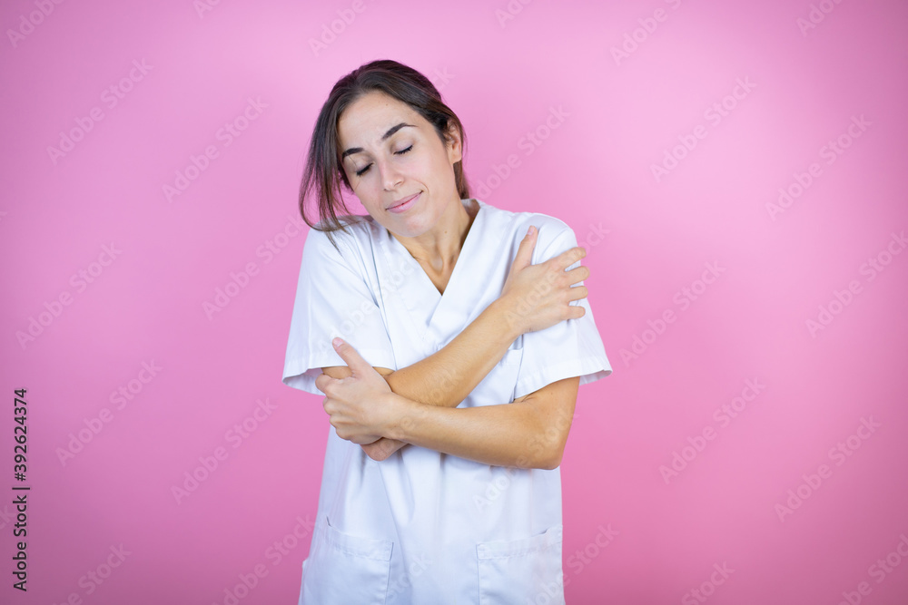 Young brunette doctor girl wearing nurse or surgeon uniform over isolated pink background hugging oneself happy and positive, smiling confident Young brunette doctor girl wearing nurse or surgeon uniform over isolated pink background hugging oneself happy and positive, smiling confident
