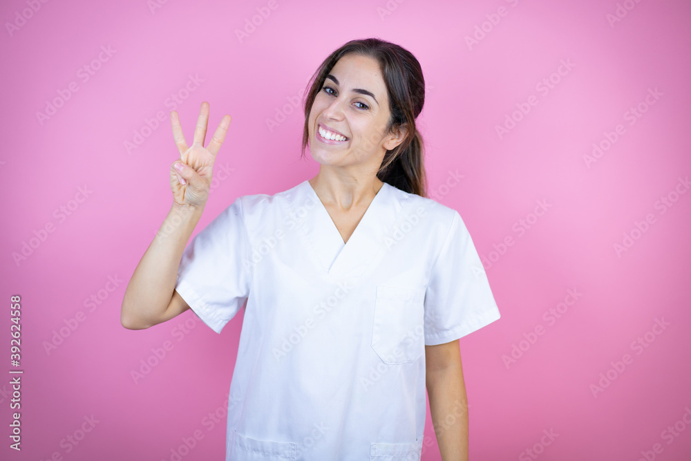 Young brunette doctor girl wearing nurse or surgeon uniform over isolated pink background showing and pointing up with fingers number three while smiling confident and happy Young brunette doctor girl wearing nurse or surgeon uniform over isolated pink background showing and pointing up with fingers number three while smiling confident and happy