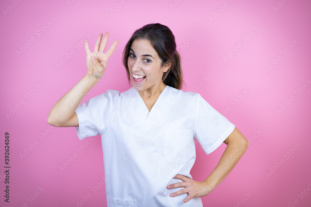 Young brunette doctor girl wearing nurse or surgeon uniform over isolated pink background showing and pointing up with fingers number four while smiling confident and happy Young brunette doctor girl wearing nurse or surgeon uniform over isolated pink background showing and pointing up with fingers number four while smiling confident and happy