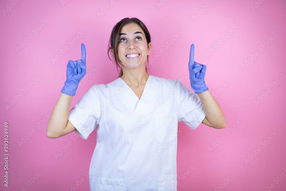Young doctor girl wearing nurse or surgeon uniform with latex gloves over isolated pink background smiling, looking up and pointing up with fingers and raised arms Young doctor girl wearing nurse or surgeon uniform with latex gloves over isolated pink background smiling, looking up and pointing up with fingers and raised arms