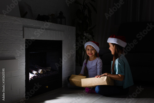 Two little sisters in pajamas having fun New Year's tree with gifts next to the fireplace © Angelov