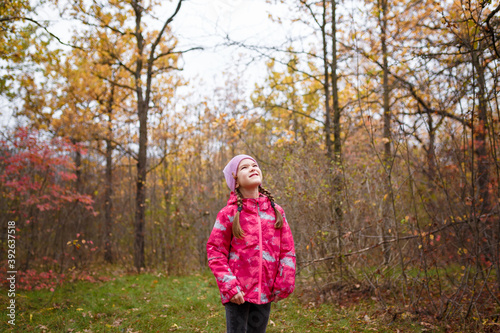 Little girl in pink jacket and knit cap standing in autumn forest