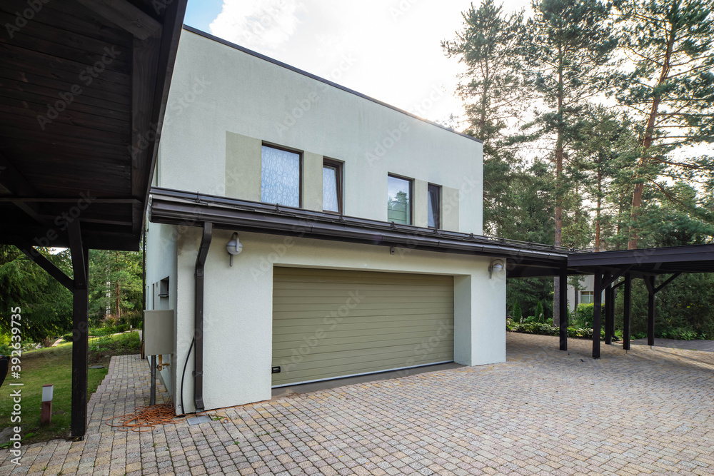 Modern exterior of luxury private house with built-in closed garage. Pines in garden. Tile. White wall with windows.