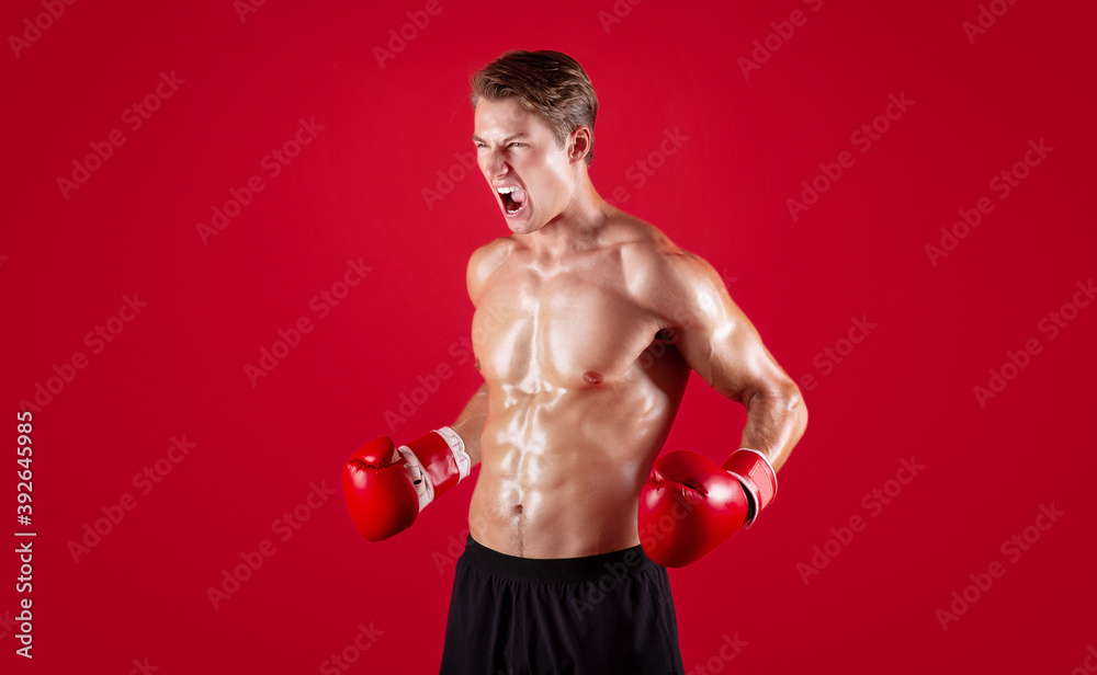 Aggressive young sportsman in boxing gloves screaming in anger on red studio background