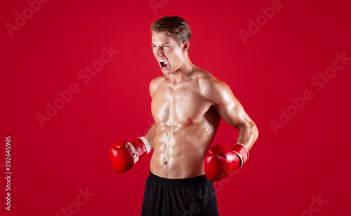 Aggressive young sportsman in boxing gloves screaming in anger on red studio background