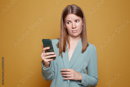 Dissatisfied lovely woman looks with disgust and aversion at modern phone, connected to high speed internet, edits photo in special app, drinks coffee to go, poses isolated on yellow studio wall.