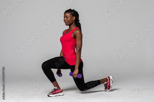 Fototapeta Naklejka Na Ścianę i Meble -  Attractive young african american sports fitness woman in sportswear posing working out isolated on gray background. Sport exercises healthy lifestyle concept. Make exercise doing lunges with dumbbell