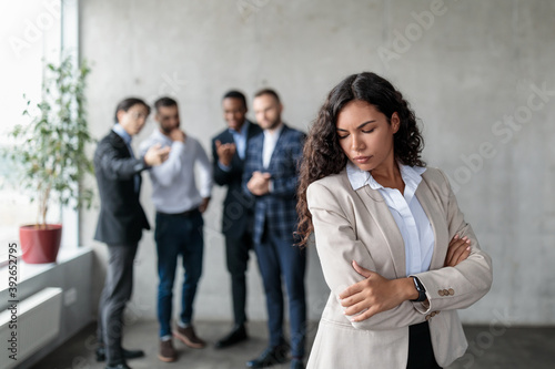 Unhappy Businesswoman Standing While Colleagues Whispering Behind Back In Office