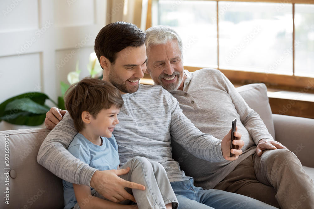 Close up happy three generations of men using smartphone together, having fun with gadget, overjoyed little boy with mature grandfather and father looking at phone screen, sitting on couch at home