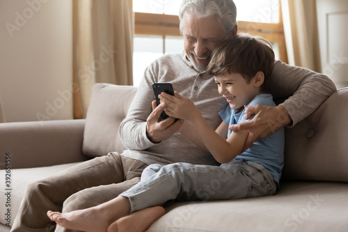Close up happy mature grandfather and grandson using phone together, smiling older man and little boy grandchild hugging, sitting on couch, looking at smartphone screen, enjoying leisure time