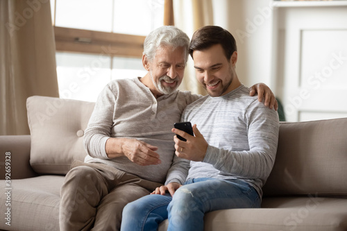 Happy older father and adult son hugging, using phone at home together, smiling mature grandfather and grandson looking at screen, browsing apps, young man teaching senior dad to use smartphone