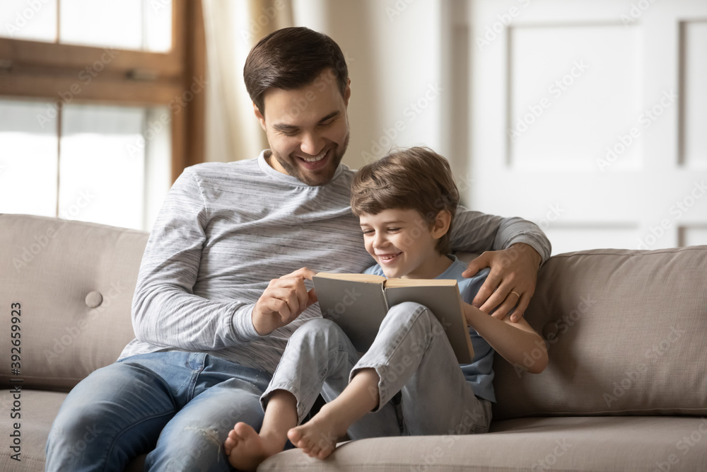 Loving happy father teaching adorable son to read, smiling dad and little boy child hugging, sitting on couch, holding book with fairy tale story, family spending weekend together, leisure time Loving happy father teaching adorable son to read, smiling dad and little boy child hugging, sitting on couch, holding book with fairy tale story, family spending weekend together, leisure time