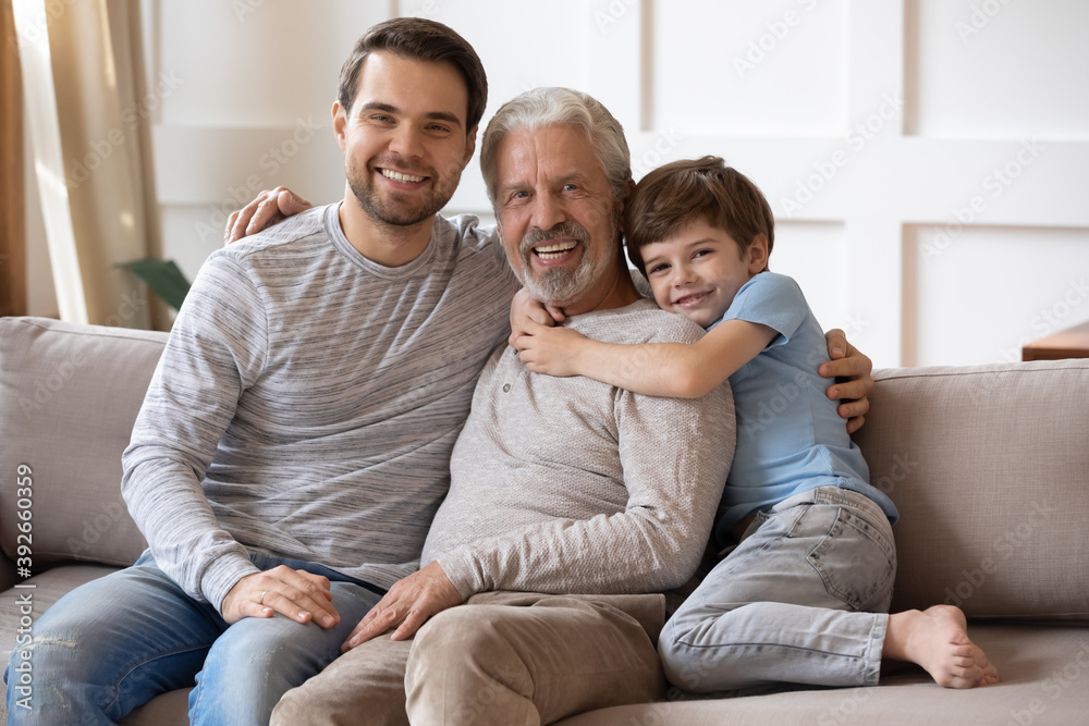 Family portrait smiling mature grandfather, young father and little boy hugging, sitting on couch at home, happy three generations of men looking at camera, posing for photo in living room Family portrait smiling mature grandfather, young father and little boy hugging, sitting on couch at home, happy three generations of men looking at camera, posing for photo in living room