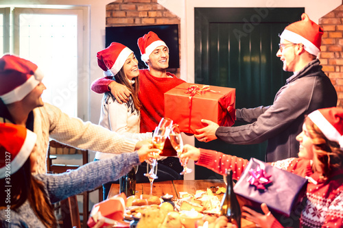 Friends with santa hat giving each other Christmas present - Champagne wine toast at house x mas dinner party - Holiday concept with people having fun together on winter time - Focus on central guy