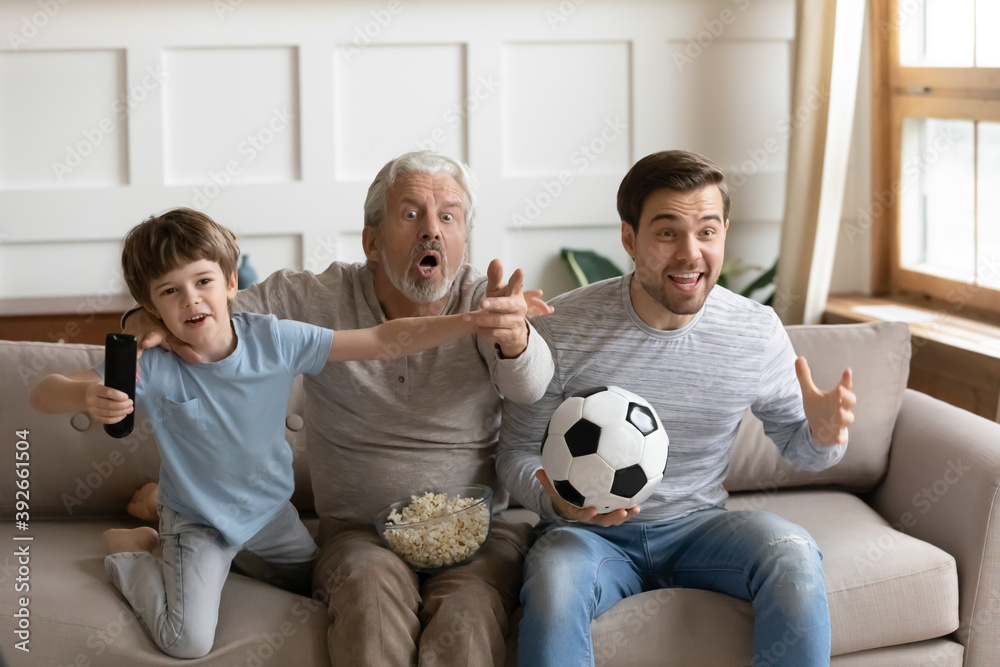 © fizkes - Overjoyed three generations of men watching match, supporting favorite football team, excited little boy with mature grandfather and father watching tv, sitting on couch with popcorn and soccer ball © fizkes - Overjoyed three generations of men watching match, supporting favorite football team, excited little boy with mature grandfather and father watching tv, sitting on couch with popcorn and soccer ball