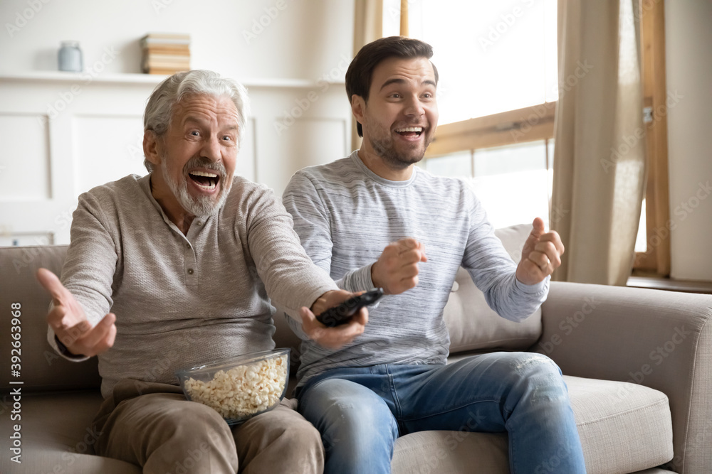 Overjoyed mature father with son watching tv, soccer match, having fun together, sitting on cozy couch with popcorn snack, excited older man with grandson supporting favorite football team Overjoyed mature father with son watching tv, soccer match, having fun together, sitting on cozy couch with popcorn snack, excited older man with grandson supporting favorite football team