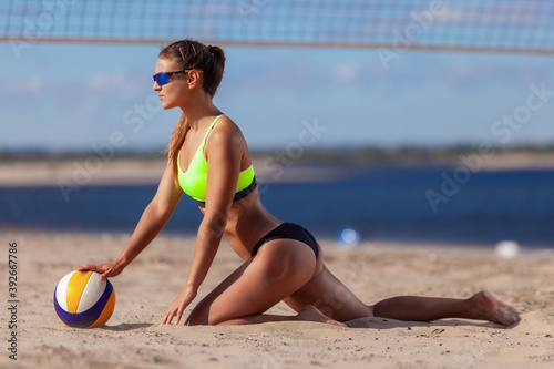 Back view of sexy athletic girl in a swimsuit and sunglasses touches a ball while sitting on the sand next to a beach volleyball net