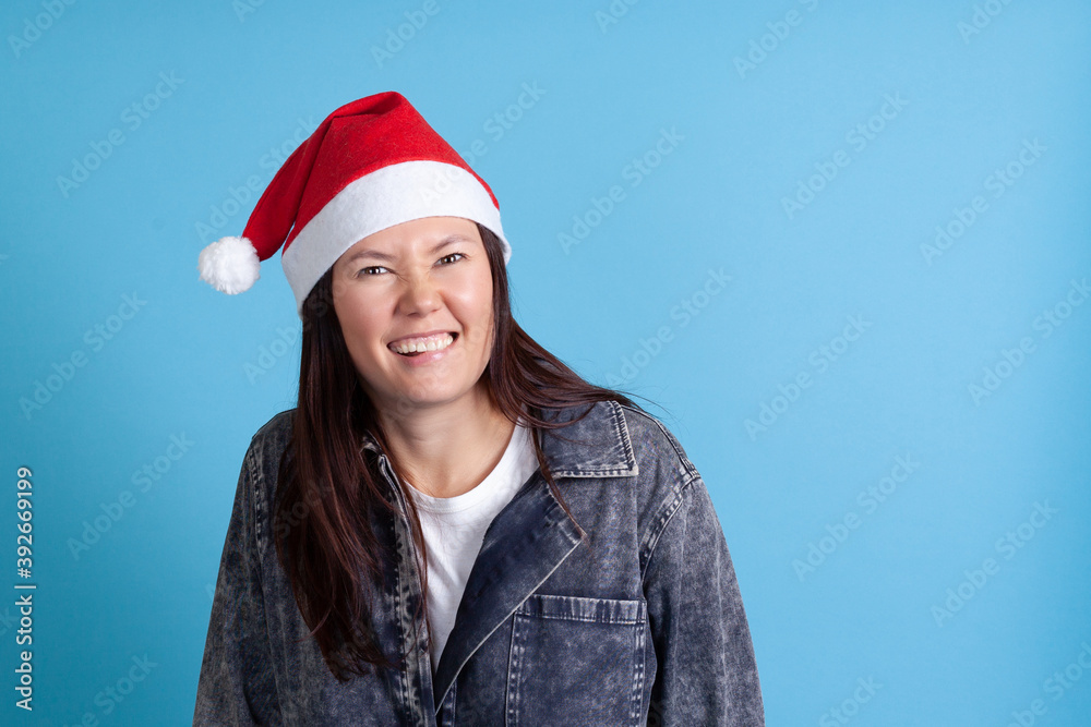 close up portrait of a mischievous Asian young woman in a Santa hat smiling and biting her lip with her teeth on a blue background. close up portrait of a mischievous Asian young woman in a Santa hat smiling and biting her lip with her teeth on a blue background.