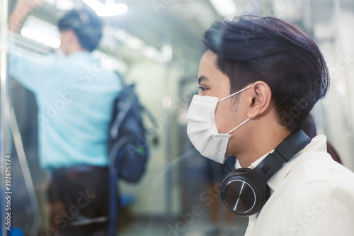 portrait young Asian man in medical face mask with headphone sitting in public metro subway. people go to work in new normal lifestyle and social distancing concept