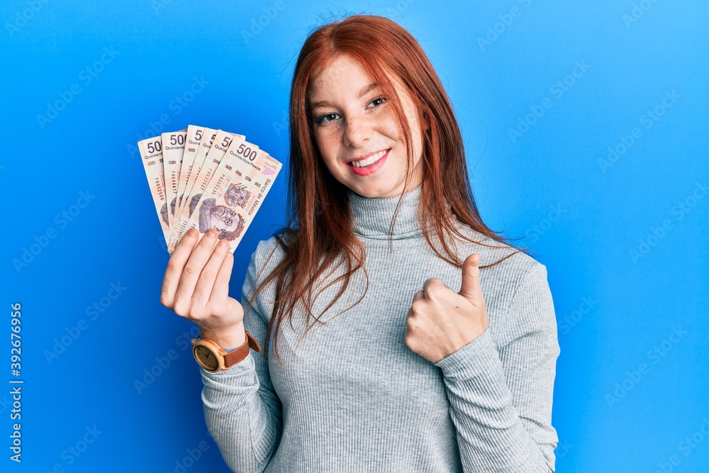 Young red head girl holding 500 mexican pesos banknotes smiling happy and positive, thumb up doing excellent and approval sign Young red head girl holding 500 mexican pesos banknotes smiling happy and positive, thumb up doing excellent and approval sign