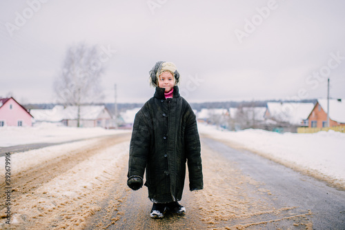 Little cheerful girl in oversized padded jacket standing on snowy road in winter day.