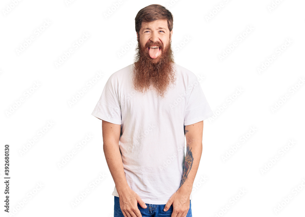 Handsome young red head man with long beard wearing casual white tshirt sticking tongue out happy with funny expression. emotion concept. Handsome young red head man with long beard wearing casual white tshirt sticking tongue out happy with funny expression. emotion concept.