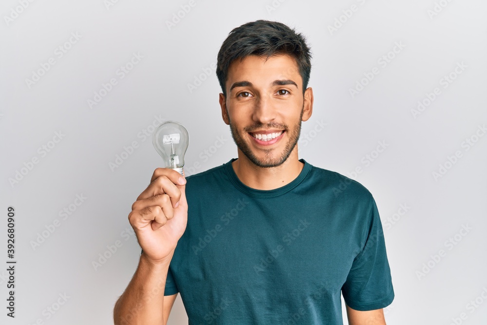 Young handsome man holding lightbulb for inspiration and idea looking positive and happy standing and smiling with a confident smile showing teeth Young handsome man holding lightbulb for inspiration and idea looking positive and happy standing and smiling with a confident smile showing teeth