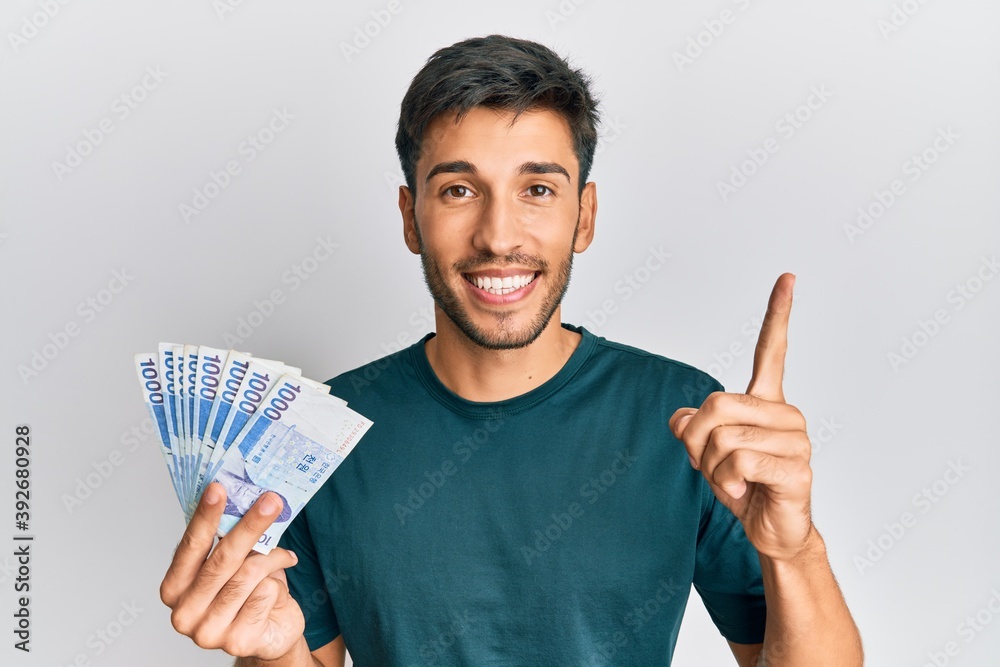 Young handsome man holding 1000 south korean won banknotes smiling with an idea or question pointing finger with happy face, number one Young handsome man holding 1000 south korean won banknotes smiling with an idea or question pointing finger with happy face, number one
