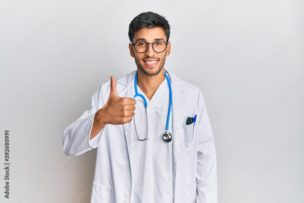 Young handsome man wearing doctor uniform and stethoscope doing happy thumbs up gesture with hand. approving expression looking at the camera showing success. Young handsome man wearing doctor uniform and stethoscope doing happy thumbs up gesture with hand. approving expression looking at the camera showing success.