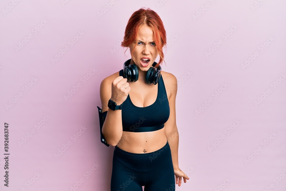 Young beautiful redhead woman wearing gym clothes and using headphones angry and mad raising fist frustrated and furious while shouting with anger. rage and aggressive concept. Young beautiful redhead woman wearing gym clothes and using headphones angry and mad raising fist frustrated and furious while shouting with anger. rage and aggressive concept.