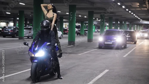 A young girl and a guy motorcyclist in a black helmet and black clothes sit on a blue motorcycle in an underground parking lot at night. Headlights of motorcycle and car shine