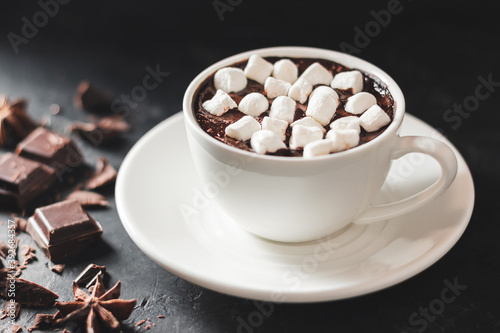 Hot chocolate drink in white cup with marshmallow, broken chocolate cubes and star anise on dark background