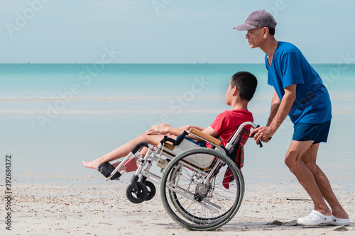 Father​ and Asian special child on wheelchair playing, doing activity on  sea beach in summer, Lifestyle of disability child, Life in the education age, Happy disabled kid in travel holidays concept.