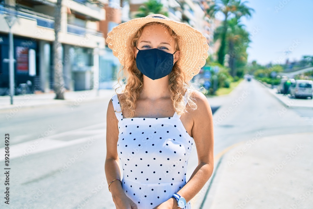 Young beautiful caucasian woman with blond hair smiling happy outdoors on a summer day wearing coronavirus safety mask