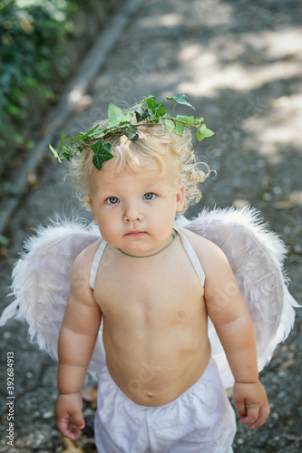 Handsome little blond curly blue eyed cupid with white feather wings and live ivy wreath. Soft focus.. Vertical photo.