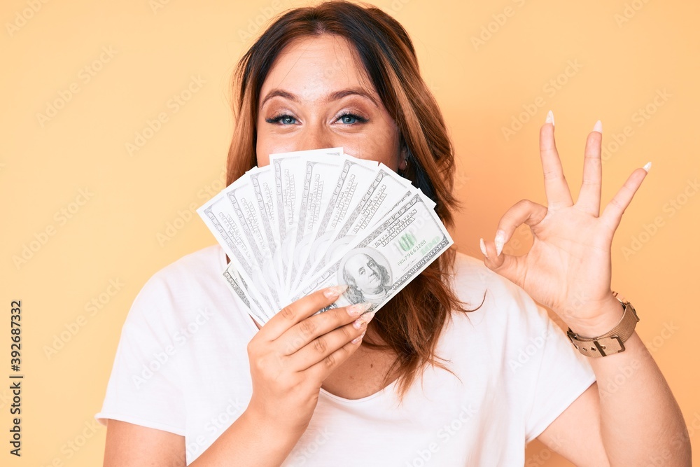 Young beautiful caucasian woman holding dollars doing ok sign with fingers, smiling friendly gesturing excellent symbol Young beautiful caucasian woman holding dollars doing ok sign with fingers, smiling friendly gesturing excellent symbol