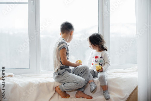 Brother and sister are sitting on the windowsill playing and eating apples. Happiness